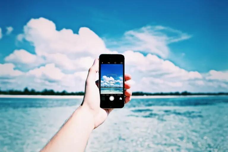 Hand holding a smartphone taking a photo of the ocean with a clear blue sky in the background.