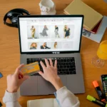 Noël Apple 2025 démarre le 12 novembre : orange, retours et zéro remise Hands holding credit card making online purchase on laptop at wooden desk with various items.
