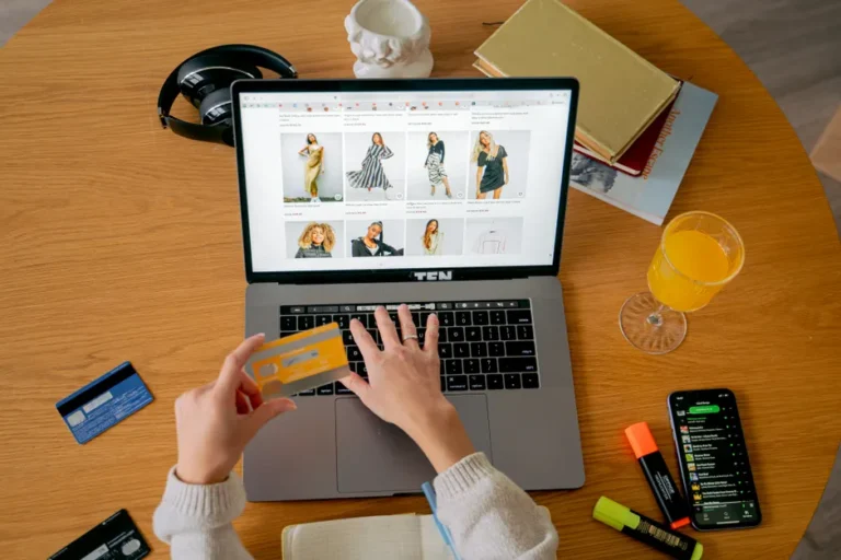 Noël Apple 2025 démarre le 12 novembre : orange, retours et zéro remise Hands holding credit card making online purchase on laptop at wooden desk with various items.