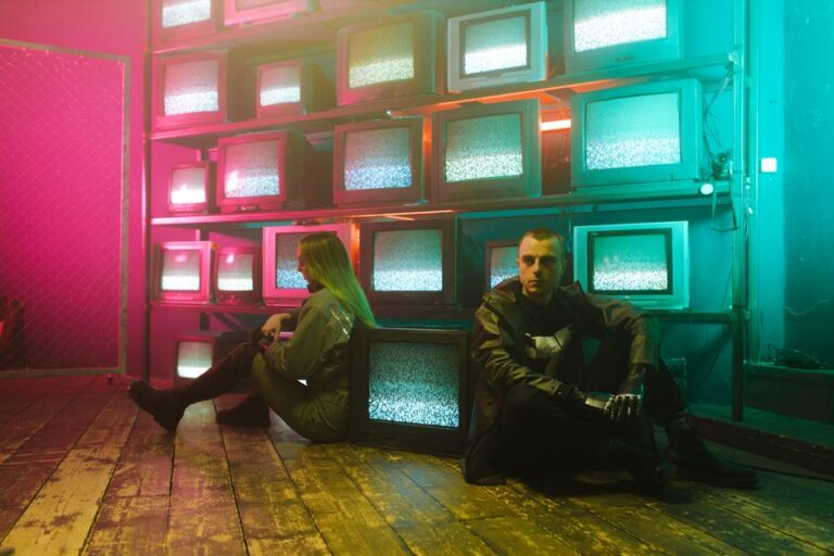 Fallen Astronaut : Apple TV aurait misé six chiffres avant la publication Two individuals in a cyberpunk setting with neon lighting and stacked television screens.