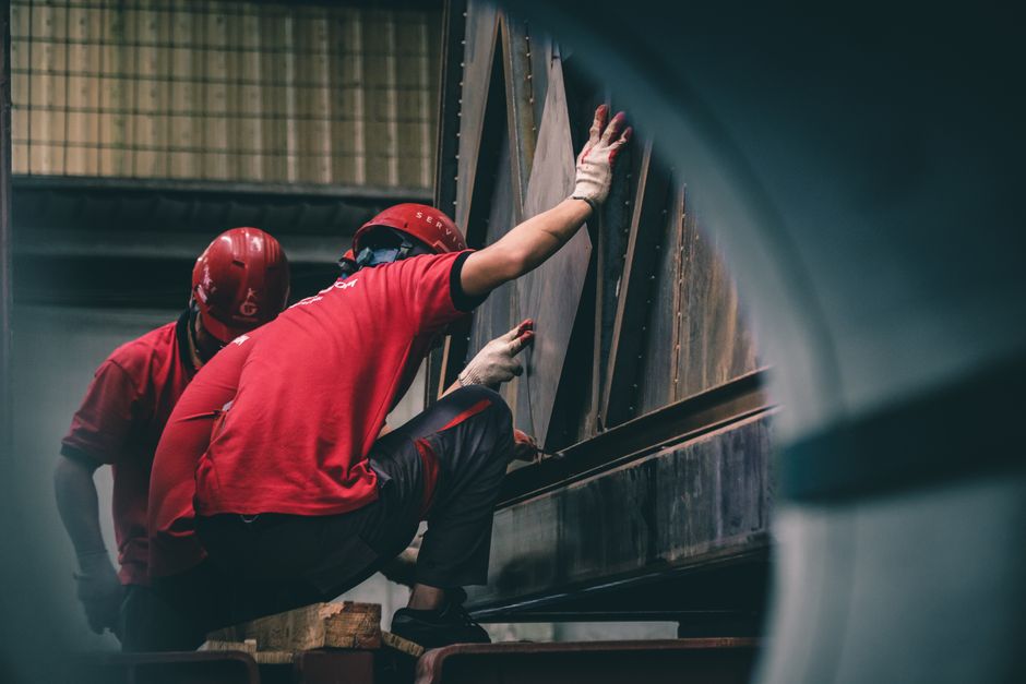 Two workers in red helmets assembling metal components inside a factory.