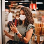 Costco Black Friday 2025 : jusqu'à 60 dollars, Apple en profite sous conditions Woman wearing mask shopping in a supermarket, checking smartphone. Captures modern retail experience.