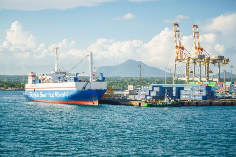 A cargo ship docked at an industrial port with shipping containers and cranes under a clear sky.