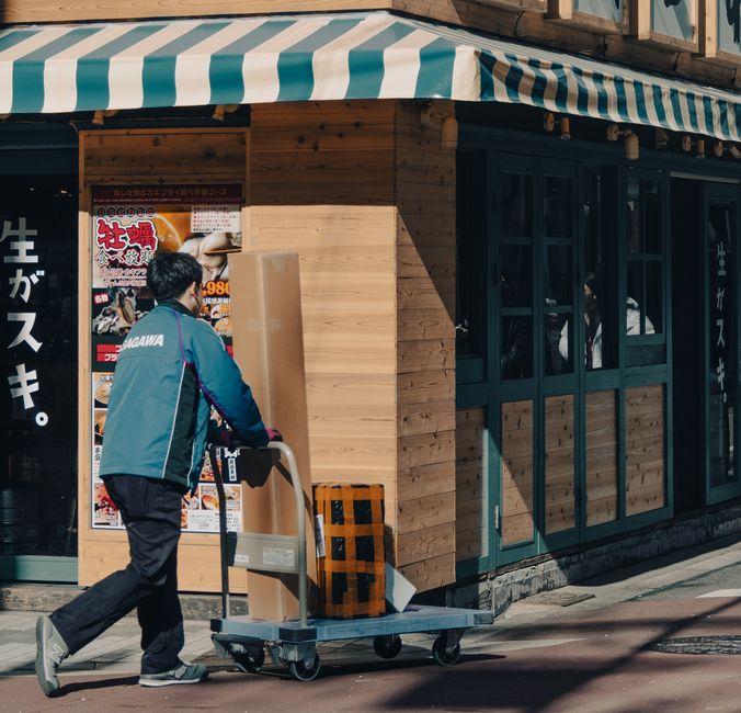 Livraison Apple en deux heures gratuite jusqu’au 24 décembre 2025 à midi, mais le stock joue les videurs A courier pushes a cart with packages outside a wooden storefront on a sunny day.