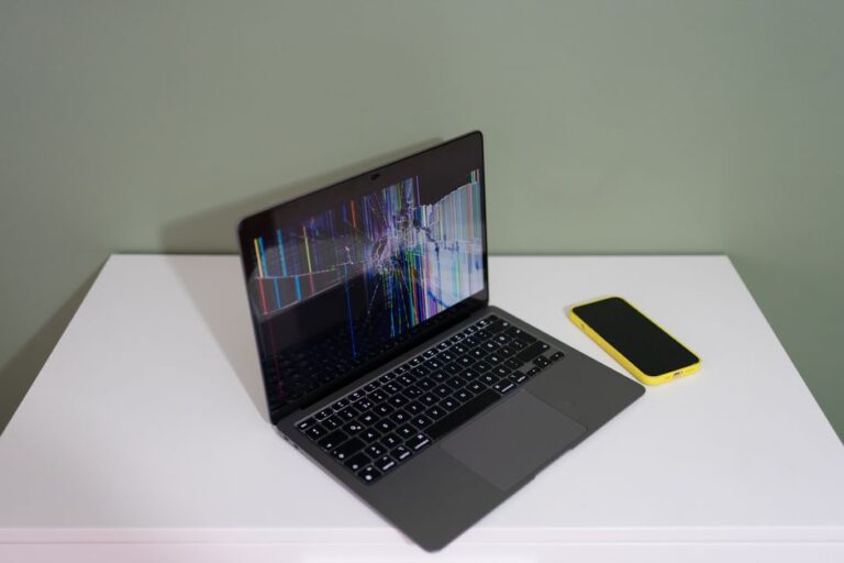 Un MacBook Air M1 encaisse un éclat d’obus en Ukraine, mais le devis pique A damaged laptop screen with colorful lines and a yellow smartphone on a white desk.
