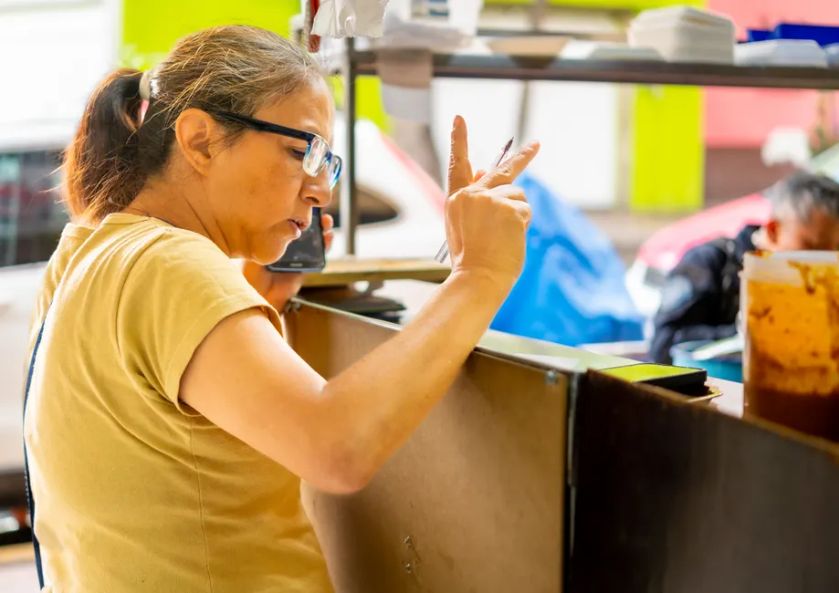 A female vendor takes an order using a smartphone at a lively outdoor food stall.