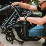 Ride1Up Portola à 1 095 dollars : pliable, mais 27 kg à porter A man folds a G-Force electric bike next to a truck, emphasizing outdoor lifestyle and convenience.