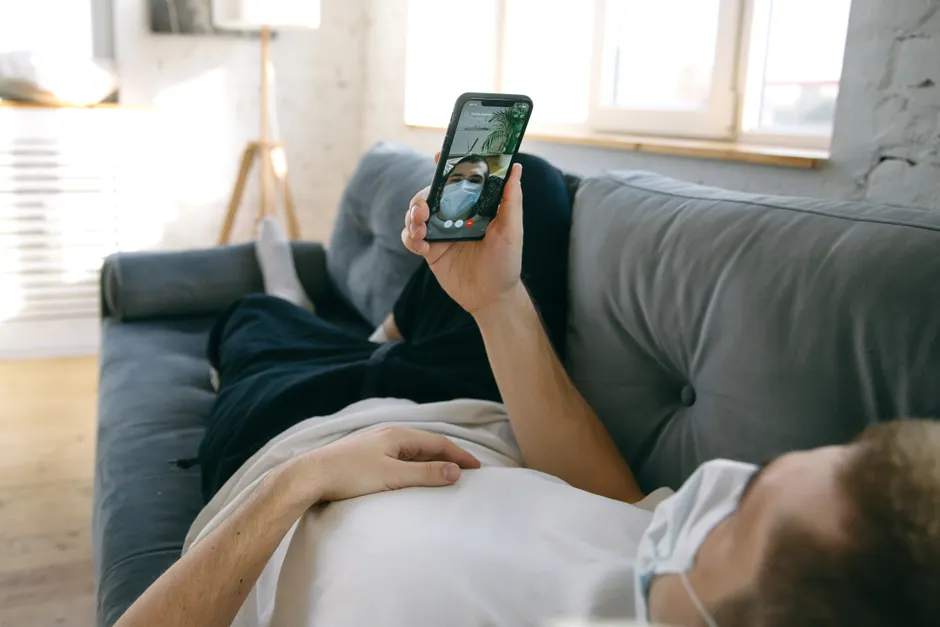 A man lying on a sofa using a smartphone for a video call while wearing a face mask indoors.
