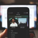 Apple Music dans ChatGPT : le DJ IA annoncé le 16 décembre 2025, sans date de sortie A person holding a smartphone with music streaming app in front of a laptop screen indoors.