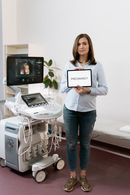 A woman holding a tablet displaying 'Pregnancy' beside an ultrasound machine in a medical clinic.