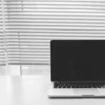 MacBook Pro M4 Max 16 pouces : 400 $ de remise, le piège des 36 ou 48 Go Black and white photo of a laptop on a white desk with window blinds in the background.