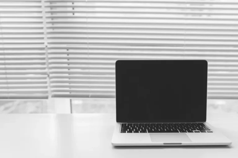 MacBook Pro M4 Max 16 pouces : 400 $ de remise, le piège des 36 ou 48 Go Black and white photo of a laptop on a white desk with window blinds in the background.