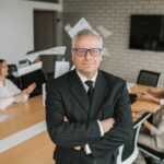 Apple 2026 : Tim Cook garde le volant, les relais se mettent en place Business executive standing confidently in meeting room with team engaged in discussion behind.