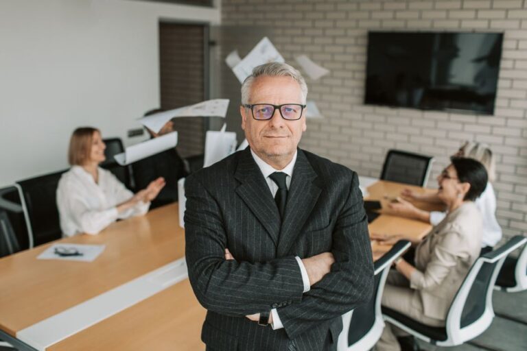 Apple 2026 : Tim Cook garde le volant, les relais se mettent en place Business executive standing confidently in meeting room with team engaged in discussion behind.