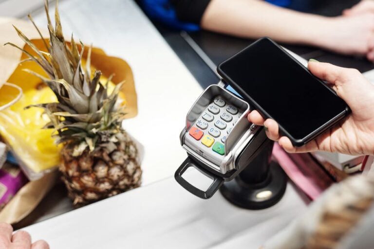 Apple Pay : 30 % chez Mattel jusqu’au 14 décembre 2025 Close-up of a customer using a smartphone for contactless payment at a retail checkout with a pineapple on the counter.