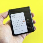 iPhone pliable en 2026 : Apple peut-elle faire plier le marché? Close-up of a hand holding a flip smartphone with a calendar on screen against a vibrant yellow background.