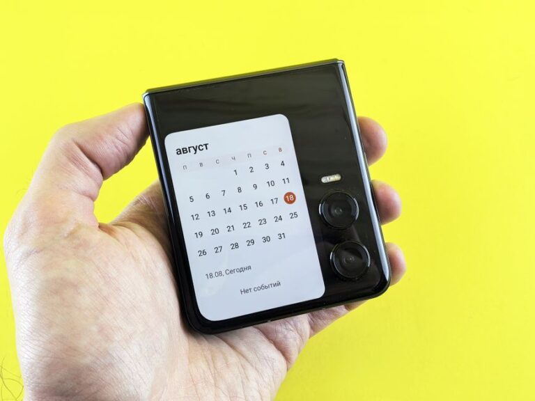 iPhone pliable en 2026 : Apple peut-elle faire plier le marché? Close-up of a hand holding a flip smartphone with a calendar on screen against a vibrant yellow background.