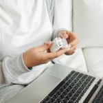 AirPods 4 ANC à 99 dollars le 1er décembre 2025: Apple tousse Close-up of hands opening wireless earbuds case near a laptop on a white couch.