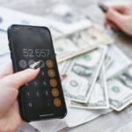 Black Friday 2025 : iPhone 17 Pro à 0 dollar chez AT&T, mais le forfait mord Close-up of hands using smartphone calculator next to dollar bills on table. Financial planning concept.