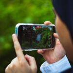 Messages par satellite arrive au Japon sur iPhone 14, le ciel en renfort Close-up of person using smartphone to film a green outdoor scene in daylight.