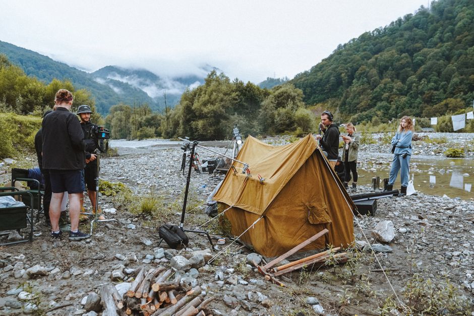 Film crew setting up camp by the river amidst forested mountains, capturing scenic landscapes.