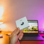AirTag Black Friday 2025 : 17,97 dollars seul, 62,99 le pack, le calcul est vite fait Hand holding an AirTag box in front of a laptop on a cozy desk setup.