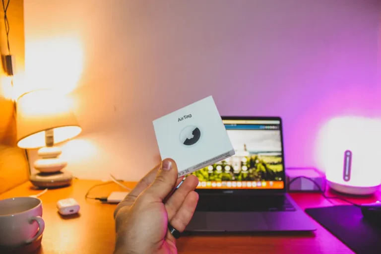 AirTag Black Friday 2025 : 17,97 dollars seul, 62,99 le pack, le calcul est vite fait Hand holding an AirTag box in front of a laptop on a cozy desk setup.