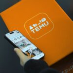 Coupon Temu Black Friday 2025 : 30 % dès 39 $, plafond à 25 $ Hand holding smartphone with Temu app beside orange Temu package.