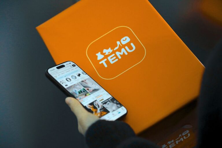 Coupon Temu Black Friday 2025 : 30 % dès 39 $, plafond à 25 $ Hand holding smartphone with Temu app beside orange Temu package.