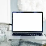 Alan Dye quitte Apple pour Meta : Liquid Glass garde le cap Image of an open laptop with a blank screen on a glass table, next to a glass of water.