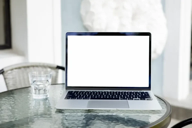 Alan Dye quitte Apple pour Meta : Liquid Glass garde le cap Image of an open laptop with a blank screen on a glass table, next to a glass of water.