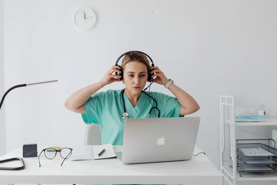 Medical professional in teal scrubs using a laptop and headset, sitting at a desk in an office setting.