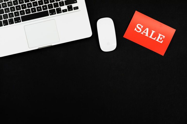 MacBook Air à 738 $ au Cyber Monday 2025 : le piège du M1 Minimalist flat lay of a laptop, mouse, and a red sale sign on black background.