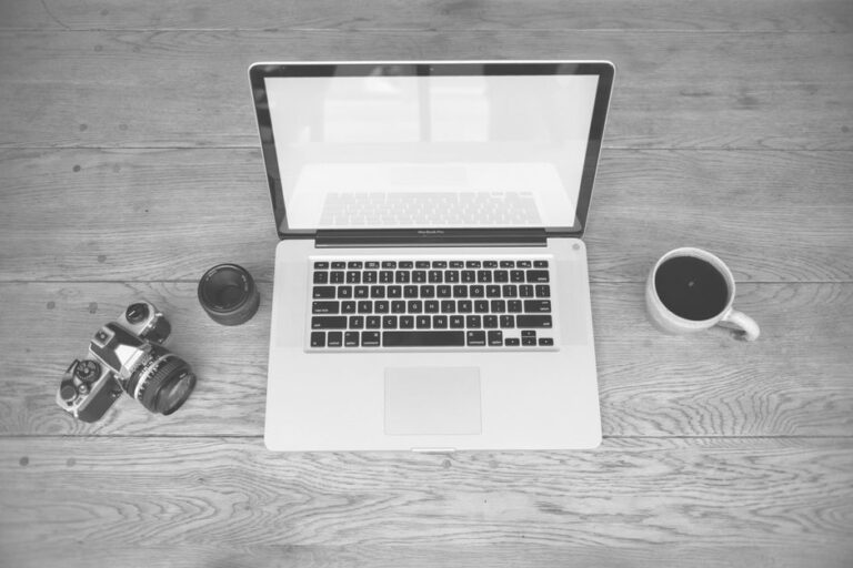 MacBook à 700 dollars en 2026 : A15 ou A18 Pro? Monochrome image of a laptop, camera, lens, and coffee cup on a wooden desk