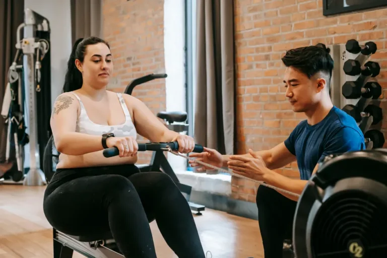Ergatta Luxe à 2 499 $ : le rameur connecté qui rame avec l’Apple Watch Positive young obese tattooed female in activewear sitting on rowing machine and doing pulling exercise during intense training with Asian male personal coach in modern gym