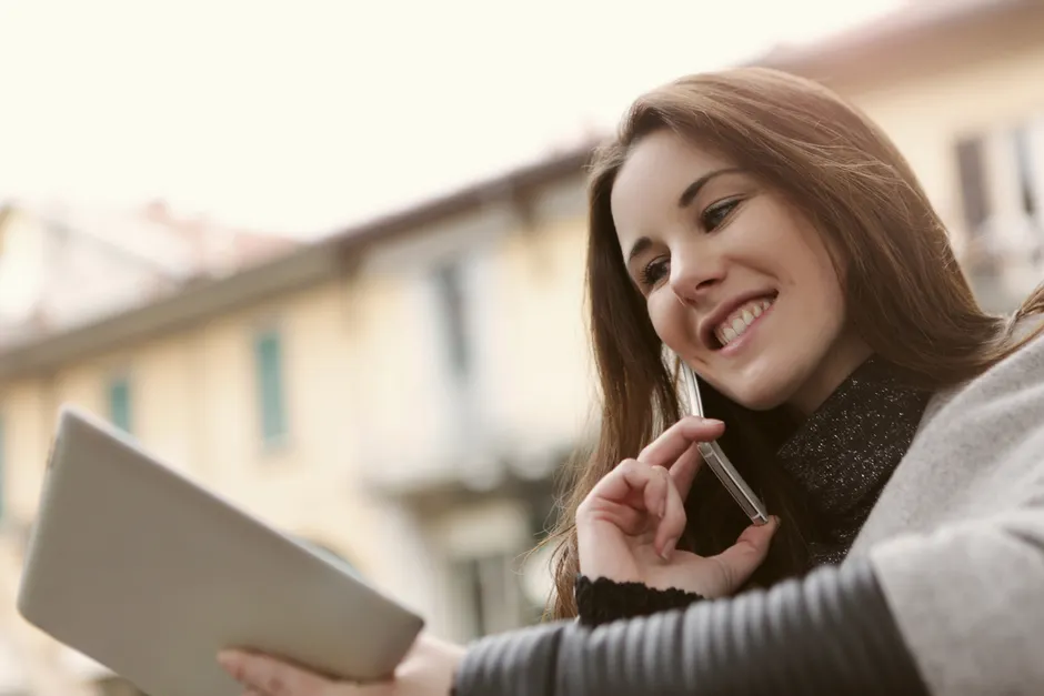 Smiling woman talking on a smartphone and holding a tablet outdoors, showcasing modern technology use.