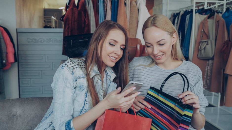 Two women enjoying a shopping spree and sharing a moment on their smartphone in a clothing store.