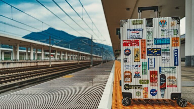 4 AirTag à 69,99 dollars chez Amazon : 17,50 dollars l’unité White suitcase adorned with travel stickers on empty railway platform, mountain backdrop.