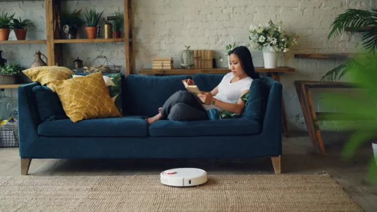 Smart home utile en 2025 : cinq appareils qui évitent l’usine à gaz Woman reading on sofa while a robot vacuum cleans the floor in a cozy living room.