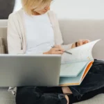 Babbel à 129,99 $ au Black Friday 2025 : le plan sans abonnement Woman sitting cross-legged on sofa, studying with a textbook and laptop, creating a cozy learning environment.