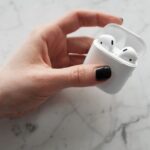 A close-up view of a hand holding white wireless earbuds in a case on a marble surface.