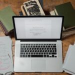A laptop surrounded by books and scripts on a wooden desk, perfect for literature and technology themes.