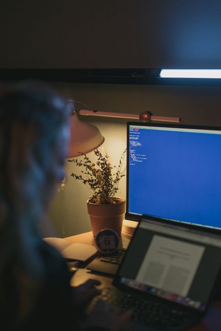A person using a computer and laptop at night with a potted plant and desk lamp.