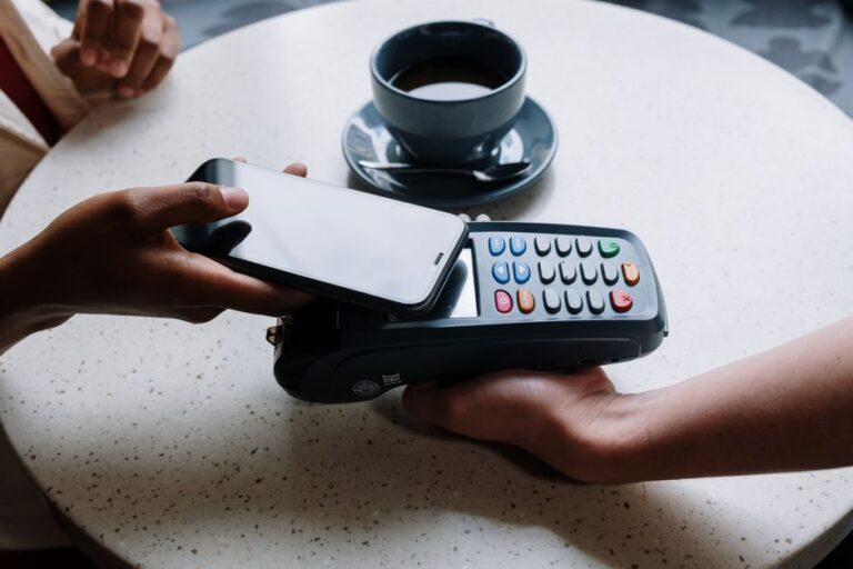 Apple Pay au Royaume-Uni : 2 milliards réclamés, mais qui paie vraiment? A person using a smartphone to make a contactless payment with a card reader on a cafe table.
