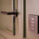 Kwikset Aura Reach à 189 dollars : Matter ouvre, Home Key reste dehors A smart light switch on a wall next to a modern door handle, emphasizing home automation.