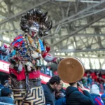 A vibrant traditional performer with feathers and drum at a lively public event indoors.