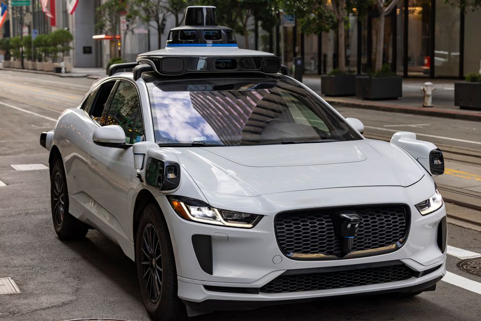 A white autonomous vehicle navigating a city street, reflecting urban architecture in daylight.