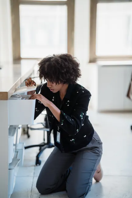 A woman kneeling and searching in an office drawer, focused on finding documents.