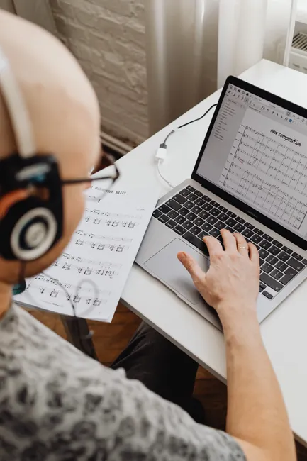 Adult musician composing music on a laptop using score sheets and headphones.