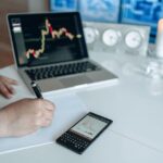 Business analyst writing with laptop and smartphone showing trend graphs on a white desk.